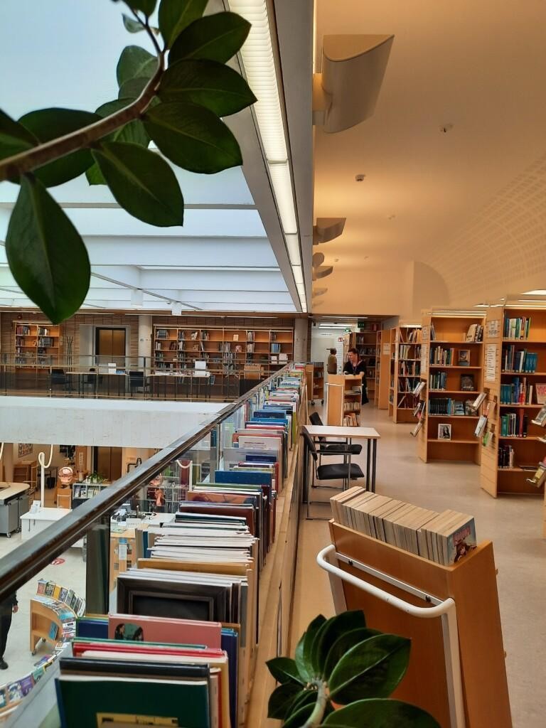 Poistokirjoja kirjaston yläkerran pitkässä hyllyssä kaiteella ja kärryissä, henkilökuntaa järjestelemässä.