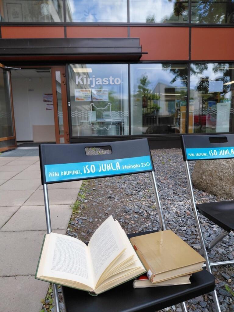 Ulkona kirjaston oven edessä kaksi tuolia joissa kaupungin juhlavuode teippaukset ja tuolilla kirjoja.